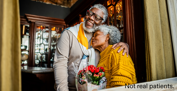 Older couple hugging, holding flowers indoors.